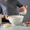Person pouring liquid from a white and pink measuring jug into a large white mixing bowl on a marble countertop with butter and eggs nearby, featured in Kitchen & Food, Cookware, Baking Supplies, Food Prep, Measuring, Best Sellers, and Promo Eligible collections.