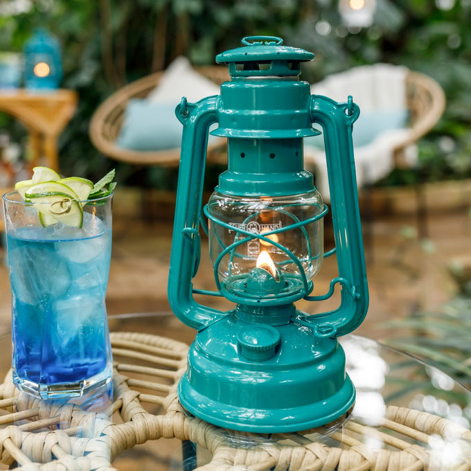 Handcrafted teal galvanized oil lantern with thermal heat-resistant globe glowing gently on a glass table in a cozy outdoor setting, ideal for Camping, Lamps & Lighting, and Emergency Supplies collections.