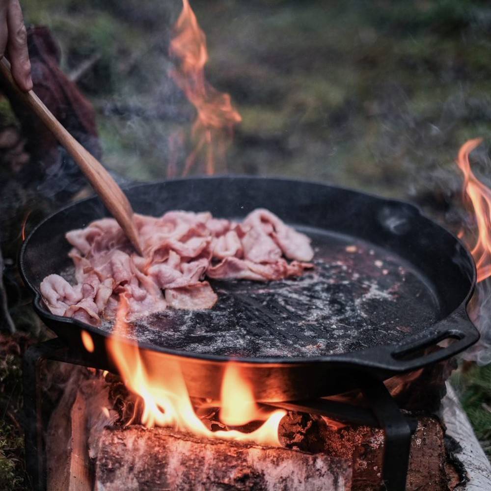 Petromax fire skillet with two side handles cooking bacon slices over an open wood fire, ideal for Farm, Garden & Tool, Camping, Kitchen & Food, Cast Iron, and Cookware collections.