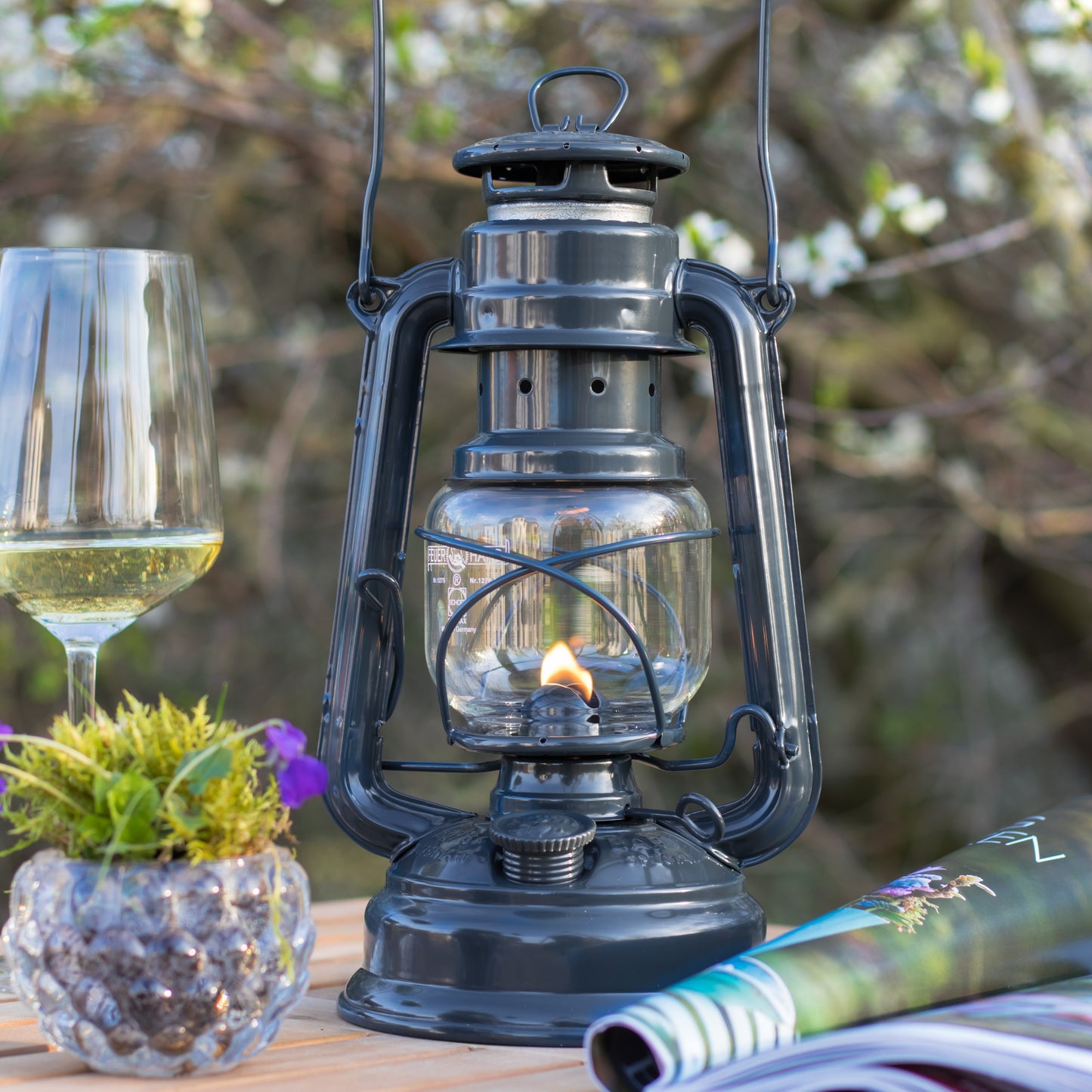 Handcrafted dark galvanized oil lantern with thermal heat-resistant globe, glowing warmly on a wooden outdoor table with a wine glass, plant, and magazine, ideal for camping and outdoor use in lamps & lighting collections.
