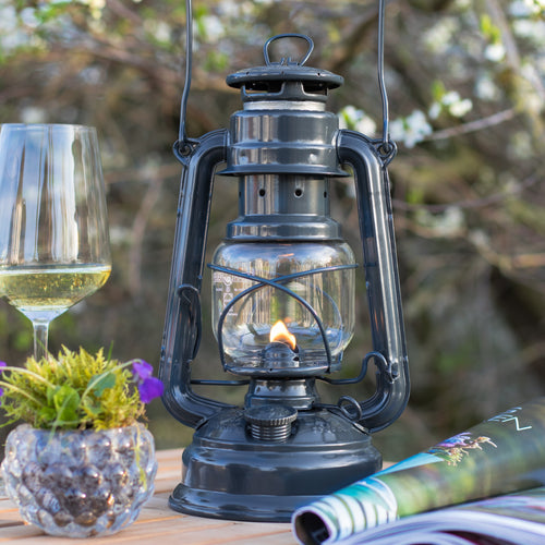 Handcrafted dark galvanized oil lantern with thermal heat-resistant globe, glowing warmly on a wooden outdoor table with a wine glass, plant, and magazine, ideal for camping and outdoor use in lamps & lighting collections.
