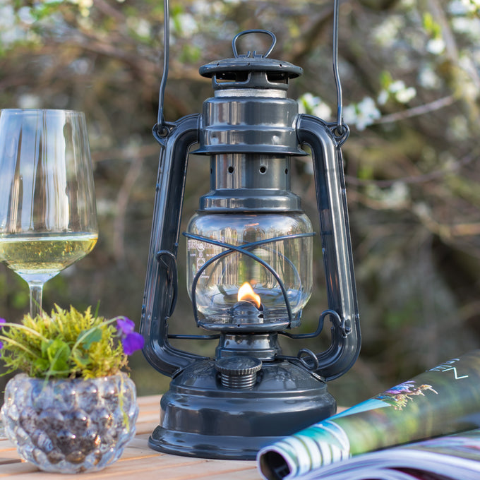 Handcrafted dark galvanized oil lantern with thermal heat-resistant globe, glowing warmly on a wooden outdoor table with a wine glass, plant, and magazine, ideal for camping and outdoor use in lamps & lighting collections.