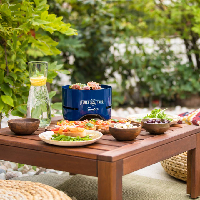Deep blue tabletop charcoal grill with food cooking on the grate, placed on a wooden outdoor table surrounded by fresh salads and olives, ideal for Farm, Garden & Tool and Camping collections.