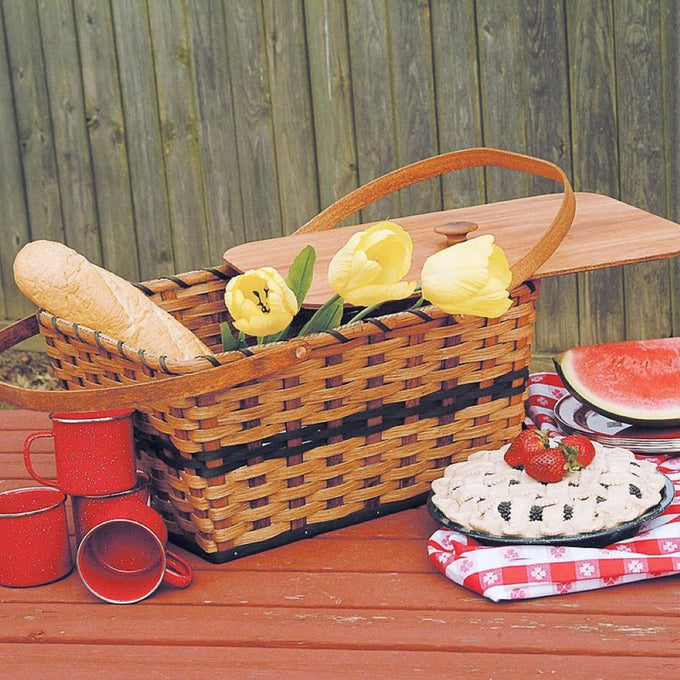 Handcrafted Amish woven basket with light-stained reeds and hunter green accents, featuring plywood lid and handles, filled with bread and yellow tulips, set for picnic in outdoor farm setting.