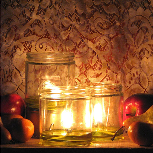 Three glowing handcrafted olive oil lamps with wide flat wicks burning warmly in clear glass jars, surrounded by fruit, featured in Lamps & Lighting, USA Made, and Olive Oil Lamps collections.
