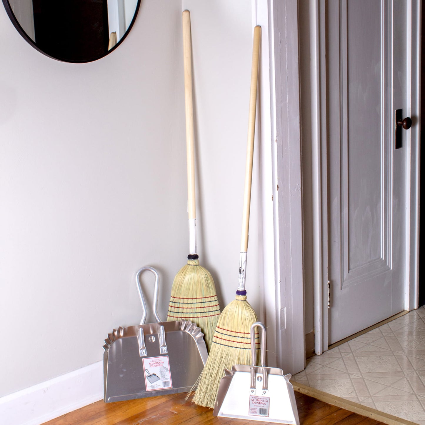 Two traditional brooms with wooden handles standing next to two sturdy metal dustpans with hanging loops, displayed in a home entryway, highlighting household cleaning utensils in USA Made and Best Sellers collections.