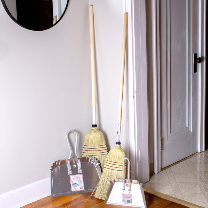 Two traditional brooms with wooden handles standing next to two sturdy metal dustpans with hanging loops, displayed in a home entryway, highlighting household cleaning utensils in USA Made and Best Sellers collections.