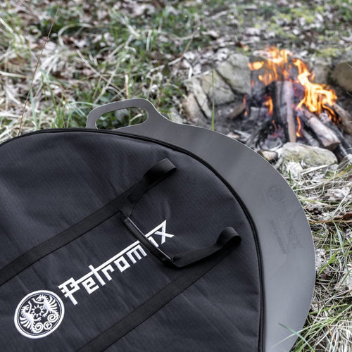 Black Petromax transport bag with sturdy handles protecting a metal griddle outdoors near a small campfire, ideal for Farm, Garden & Tool, Camping, Best Sellers, and Promo Eligible collections.