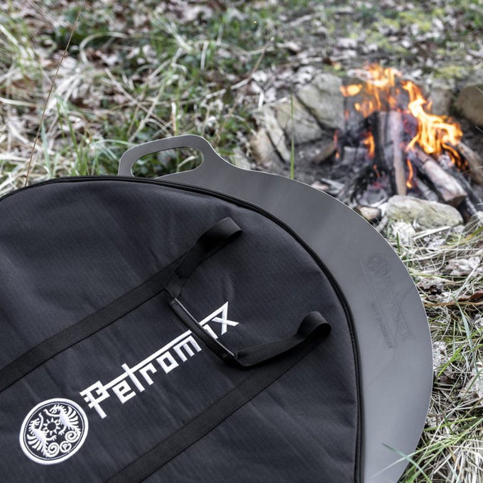 Black Petromax transport bag with sturdy handles protecting a metal griddle outdoors near a small campfire, ideal for Farm, Garden & Tool, Camping, Best Sellers, and Promo Eligible collections.