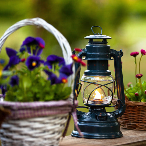 Handcrafted dark galvanized oil lantern with thermal heat-resistant SUPRAX globe, glowing warmly on a wooden table amid outdoor flower baskets, ideal for camping and emergency supplies in best sellers and promo collections.
