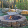 Outdoor fire pit constructed from stacked stone blocks with a stainless steel adjustable grill grate suspended above burning wood, ideal for camping and backyard grilling.