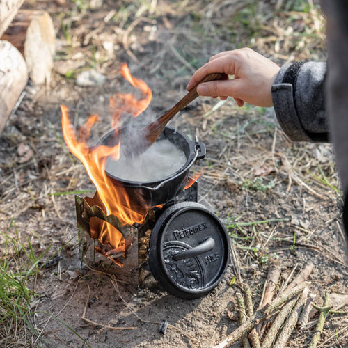 Small cast iron Dutch oven with legs cooking over an open campfire outdoors, steam rising as a hand stirs with a wooden spoon, ideal for Kitchen & Food, Cookware, Cast Iron collections.