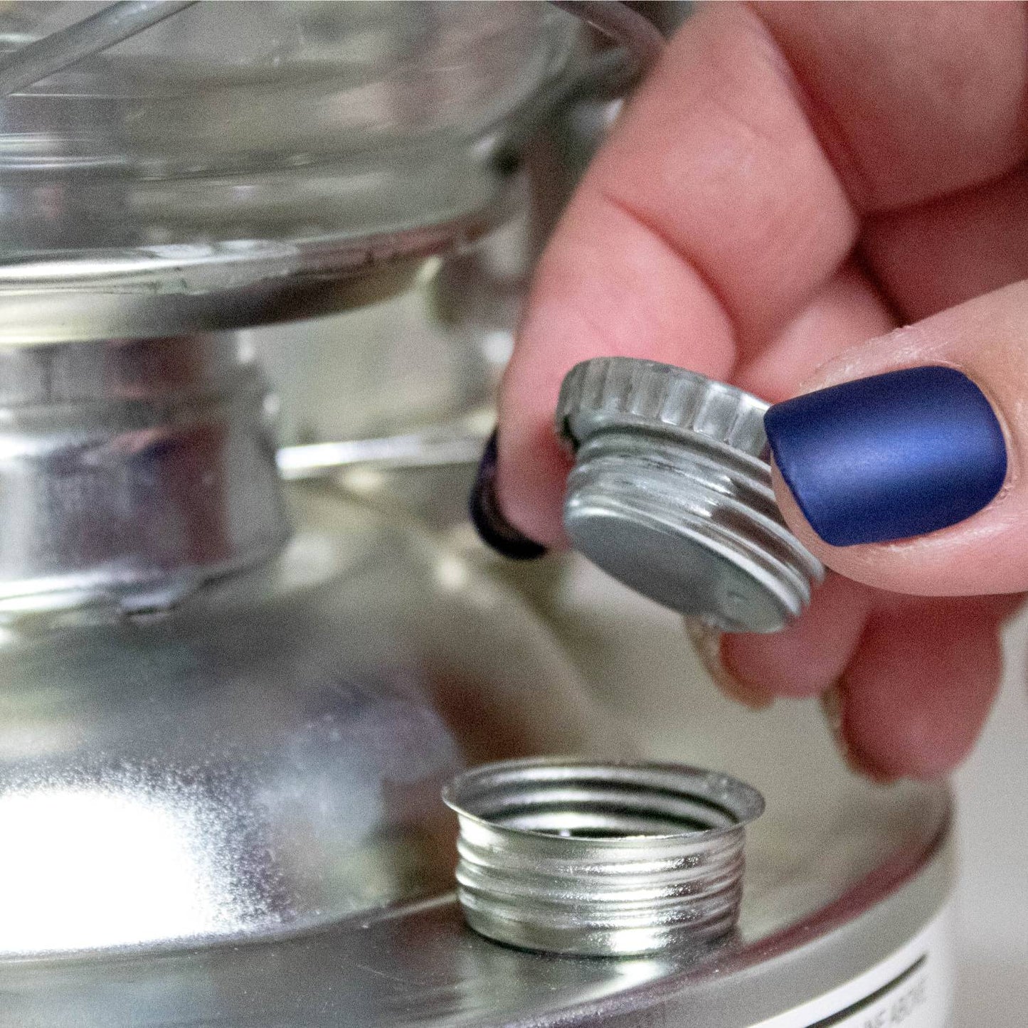 Close-up of a hand with blue matte nail polish removing a silver metal fuel cap from a handcrafted oil lantern, featured in Farm, Garden & Tool, Camping, and Emergency Supplies collections.