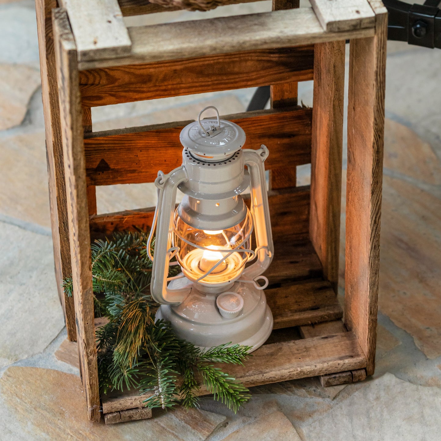 Handcrafted galvanized oil lantern with thermal heat-resistant globe glowing warmly inside a rustic wooden crate, surrounded by evergreen branches, ideal for Camping and Emergency Supplies collections.