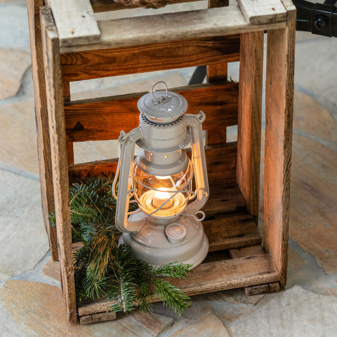 Handcrafted galvanized oil lantern with thermal heat-resistant globe glowing warmly inside a rustic wooden crate, surrounded by evergreen branches, ideal for Camping and Emergency Supplies collections.