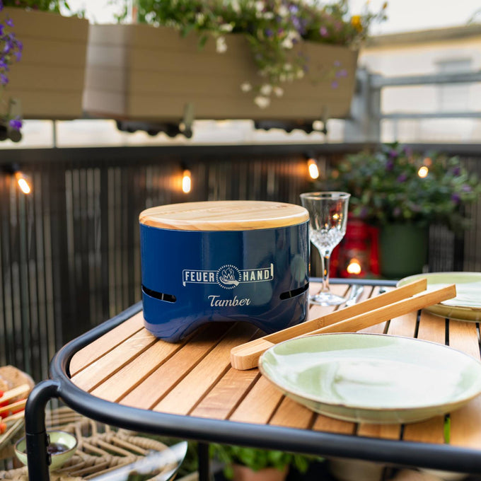 Compact handcrafted blue tabletop charcoal grill with a wooden lid, placed on a round wooden outdoor table set for dining, perfect for Farm, Garden & Tool and Camping collections.
