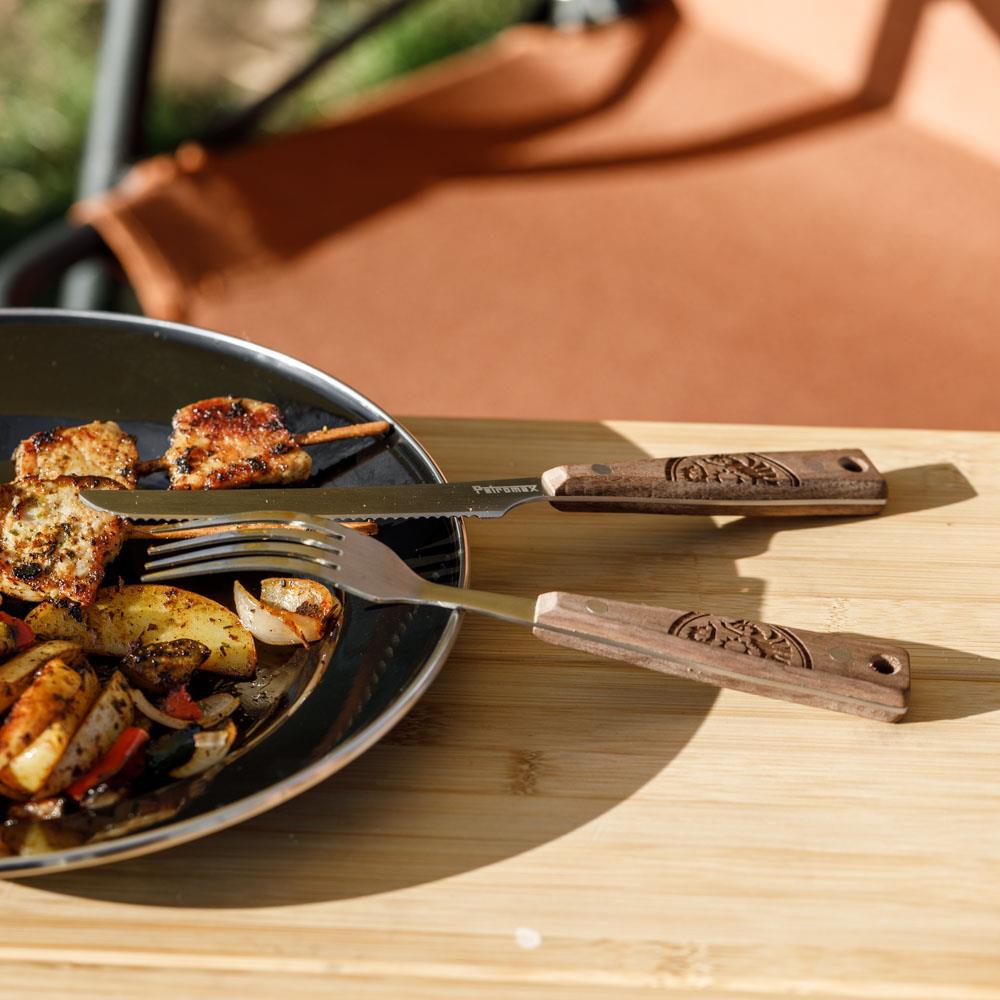 Camping cutlery set featuring a serrated knife and fork with engraved wooden handles resting on a wooden surface next to a plate of grilled vegetables and meat, suitable for Farm, Garden & Tool, Kitchen & Food, Camping, and Dinnerware collections.