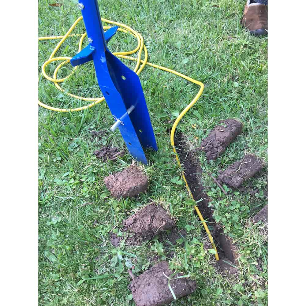 Blue trenching shovel with narrow blade cutting a clean trench in grassy soil, surrounded by removed soil clumps and a yellow cable, suitable for farm tools and garden digging tasks.