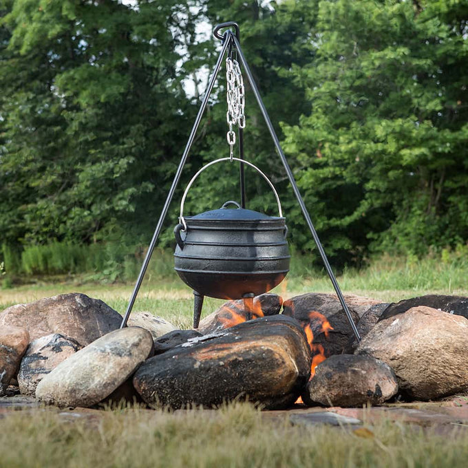 Handcrafted cast iron kettle with a rounded body and fitted lid, suspended from a tripod above a campfire surrounded by stones, shown in an outdoor Farm, Garden & Tool, Camping setting.