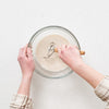 Person mixing liquid ingredients in a clear glass bowl using a wooden-handled whisk, demonstrating a step in sourdough baking for Kitchen & Food, Baking Supplies, and Cookware collections.