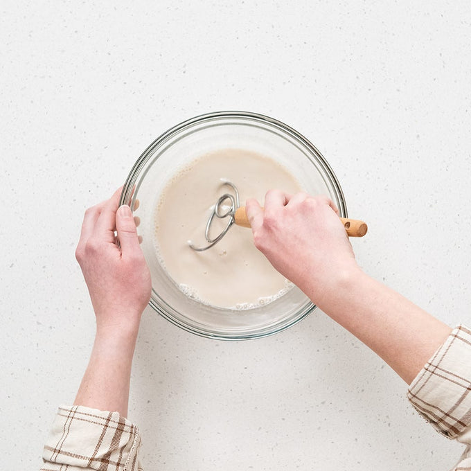 Person mixing liquid ingredients in a clear glass bowl using a wooden-handled whisk, demonstrating a step in sourdough baking for Kitchen & Food, Baking Supplies, and Cookware collections.