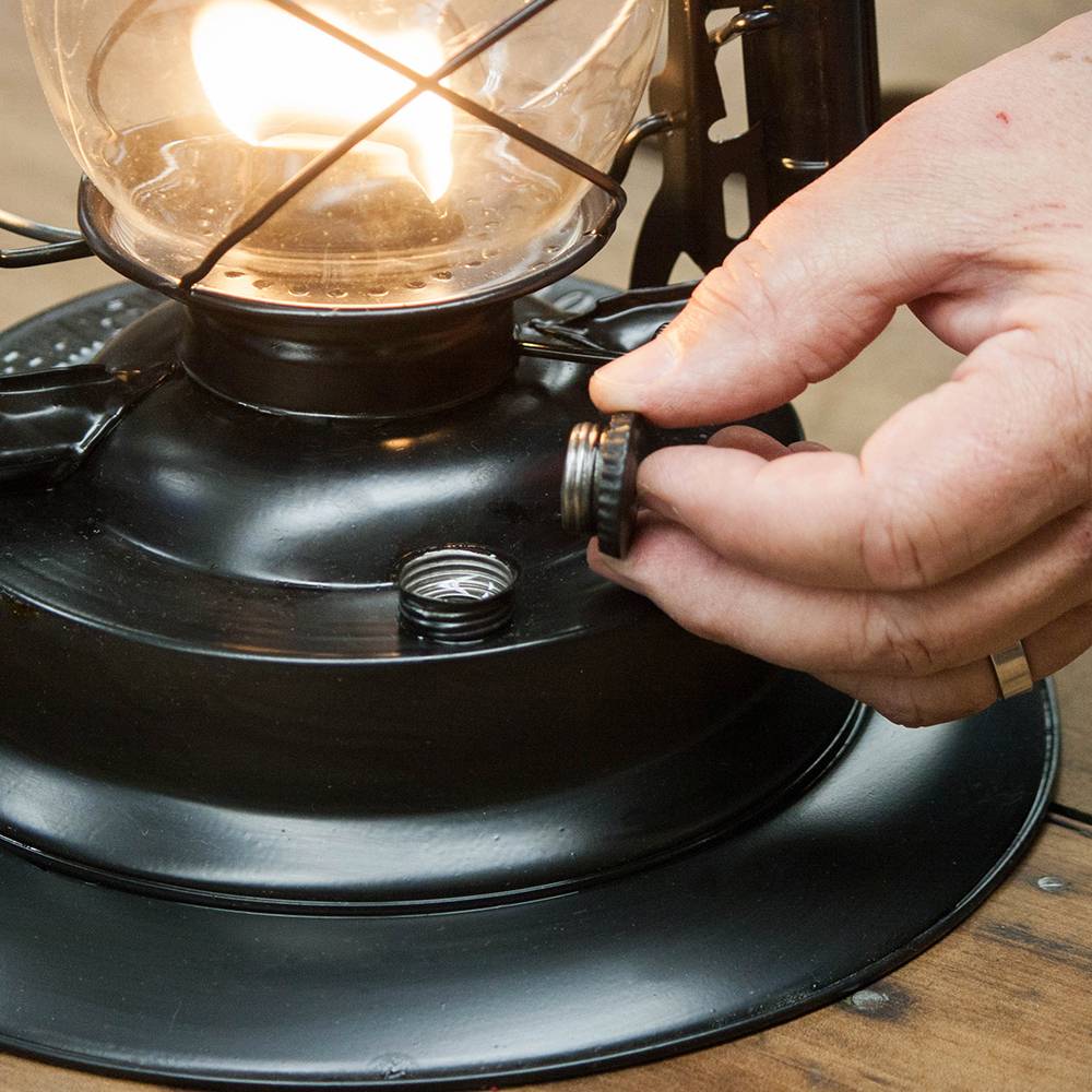 Handcrafted black powder-coated steel lantern with lit flame and user adjusting fuel cap, designed for reliable light, heat, and cooking in camping, emergency, and outdoor use.