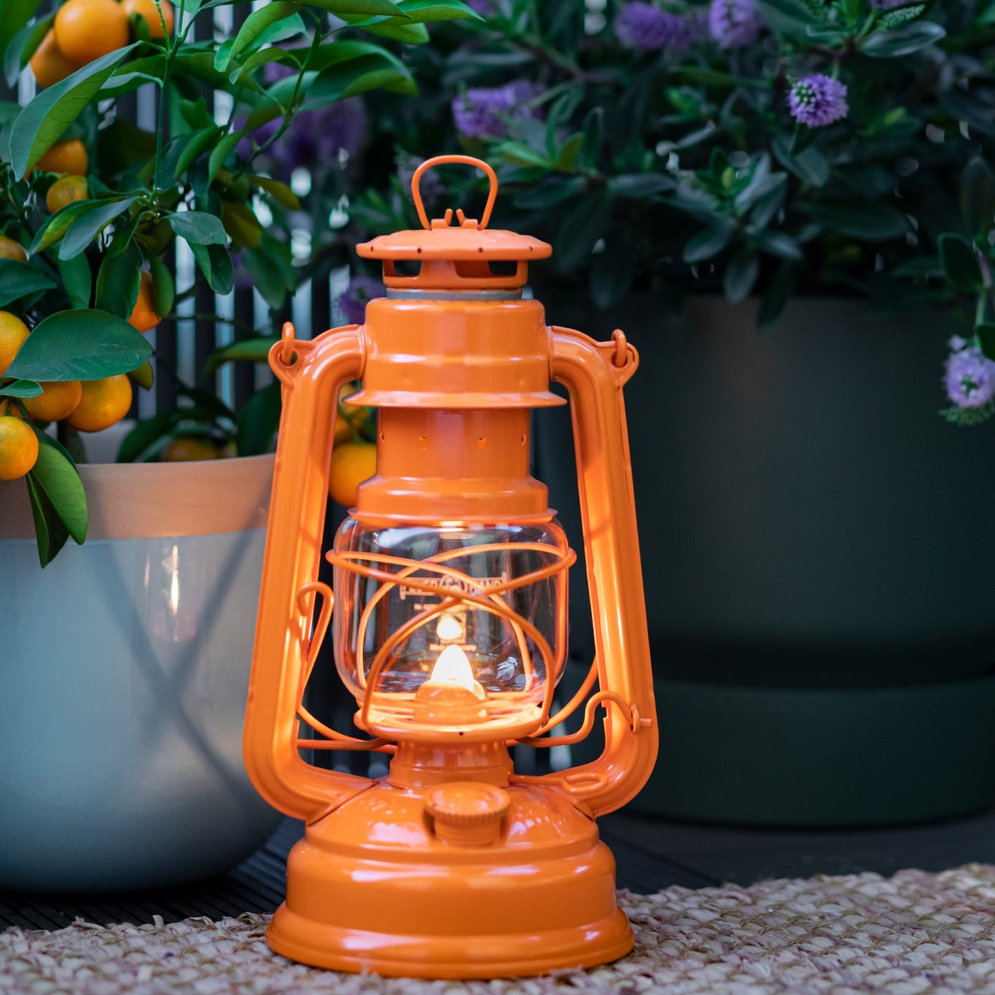 Handcrafted bright orange galvanized oil lantern with thermal heat-resistant globe glowing gently, poised on outdoor patio beside potted plants, perfect for Camping, Lamps & Lighting, and Emergency Supplies collections.