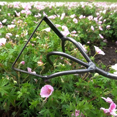Close-up of a handcrafted metal garden tool with a triangular blade and looped support, positioned among green foliage and small pink flowers in a garden setting, featured in Farm, Garden & Tool and Garden Accessories collections.