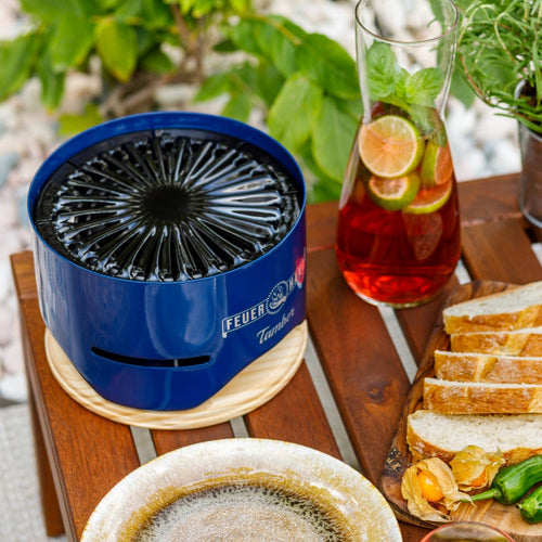 Compact blue tabletop charcoal grill with a detailed radial grate, set on a wooden trivet beside a pitcher of citrus beverage and sliced bread, ideal for Farm, Garden & Tool and Grilling collections.