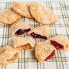 Assortment of handcrafted, Amish-style fry pies with thick golden crusts and fruit fillings like cherry, blueberry, and apple on a green and white checkered cloth, ideal for Kitchen & Food, Snacks and Sweets, Holiday Hosting, USA Made, Best Sellers, and Promo Eligible collections.