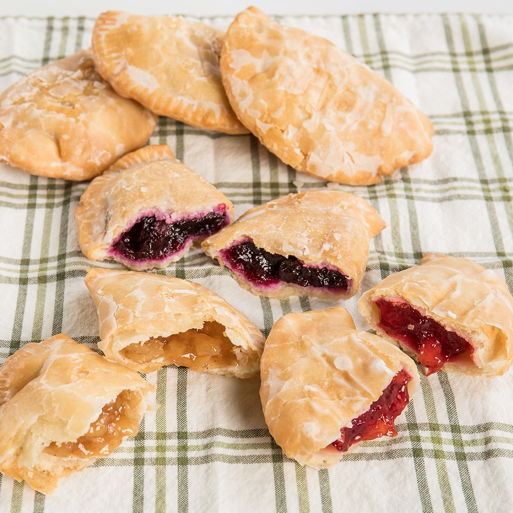 Assortment of handcrafted, Amish-style fry pies with thick golden crusts and fruit fillings like cherry, blueberry, and apple on a green and white checkered cloth, ideal for Kitchen & Food, Snacks and Sweets, Holiday Hosting, USA Made, Best Sellers, and Promo Eligible collections.