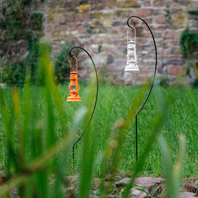 Two black metal lantern holders with curved hooks suspending an orange and a white lantern outdoors in a grassy garden near a stone wall, crafted for lamps & lighting, camping, and emergency supplies, featuring a handcrafted, two-piece design for easy assembly and transport.