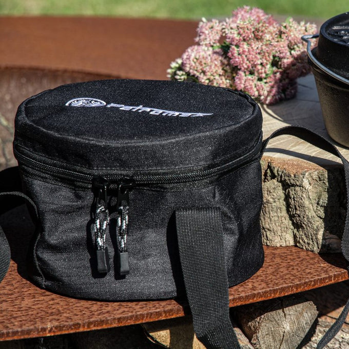 Round black ripstop fabric case with dual zipper and shoulder strap, designed for protecting and carrying Dutch ovens, shown outdoors with wood and flowers, suited for Farm, Garden & Tool and Camping collections.