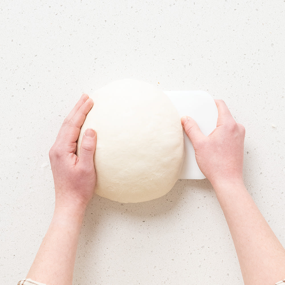 Pair of hands shaping a smooth round sourdough dough ball on a speckled white surface using a white dough scraper, ideal for Kitchen & Food, Baking Supplies, and Bread Making collections.