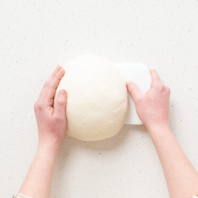 Pair of hands shaping a smooth round sourdough dough ball on a speckled white surface using a white dough scraper, ideal for Kitchen & Food, Baking Supplies, and Bread Making collections.