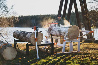 Man operating a robust chain sawmill cutting large logs outdoors near a lakeside bridge, featured in Farm, Garden & Tool, Wood Cutting and Hauling, and Best Sellers collections