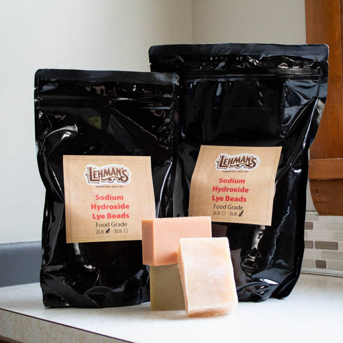Two black resealable bags of food-grade sodium hydroxide lye beads in 2-lb and 5-lb sizes with three handcrafted soap bars on a kitchen countertop, featured in Health & Wellness, Soaps and Soapmaking, Soapmaking Supplies, USA Made, and Best Sellers collections.