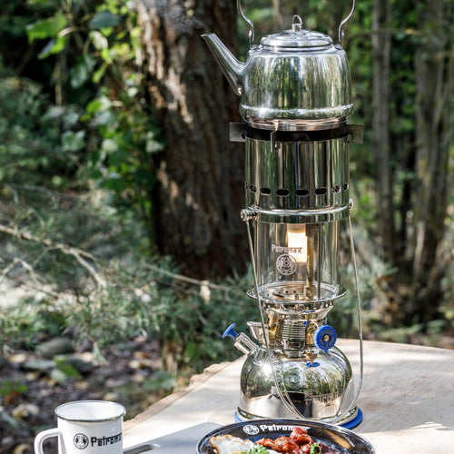 Shiny metal kettle heating on a portable cooking device adapted from a classic lamp, set outdoors on a wooden table with camping gear from Farm, Garden & Tool and Camping collections.