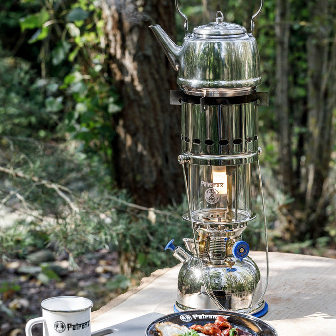 Shiny metal kettle heating on a portable cooking device adapted from a classic lamp, set outdoors on a wooden table with camping gear from Farm, Garden & Tool and Camping collections.