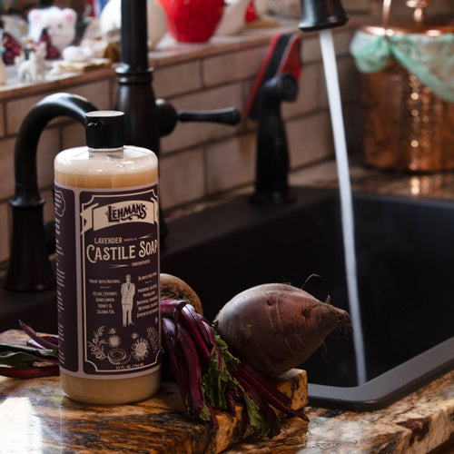 Amber bottle of Lehman's Lavender Castile soap near fresh root vegetables on a kitchen counter next to a running faucet, highlighting handcrafted, multi-use, USA made soap for Health & Wellness and Grooming collections.