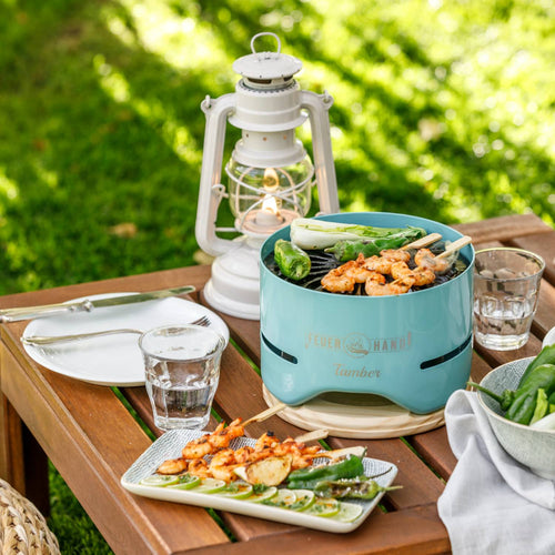 Unique handcrafted turquoise tabletop charcoal grill cooking skewers and vegetables, displayed on a wooden patio table with glassware and a vintage lantern, ideal for Farm, Garden & Tool, Camping, and Grilling collections.