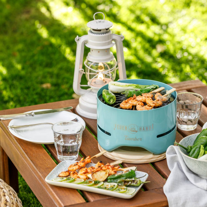 Unique handcrafted turquoise tabletop charcoal grill cooking skewers and vegetables, displayed on a wooden patio table with glassware and a vintage lantern, ideal for Farm, Garden & Tool, Camping, and Grilling collections.