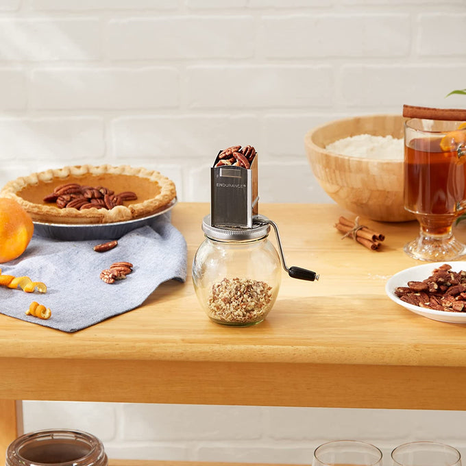 Vintage-style nut grinder with stainless steel hopper filled with pecans, glass jar beneath catching chopped nuts, displayed on wooden table with pie and baking ingredients, ideal for Kitchen & Food, Food Prep, and Cooking and Baking Helpers collections.