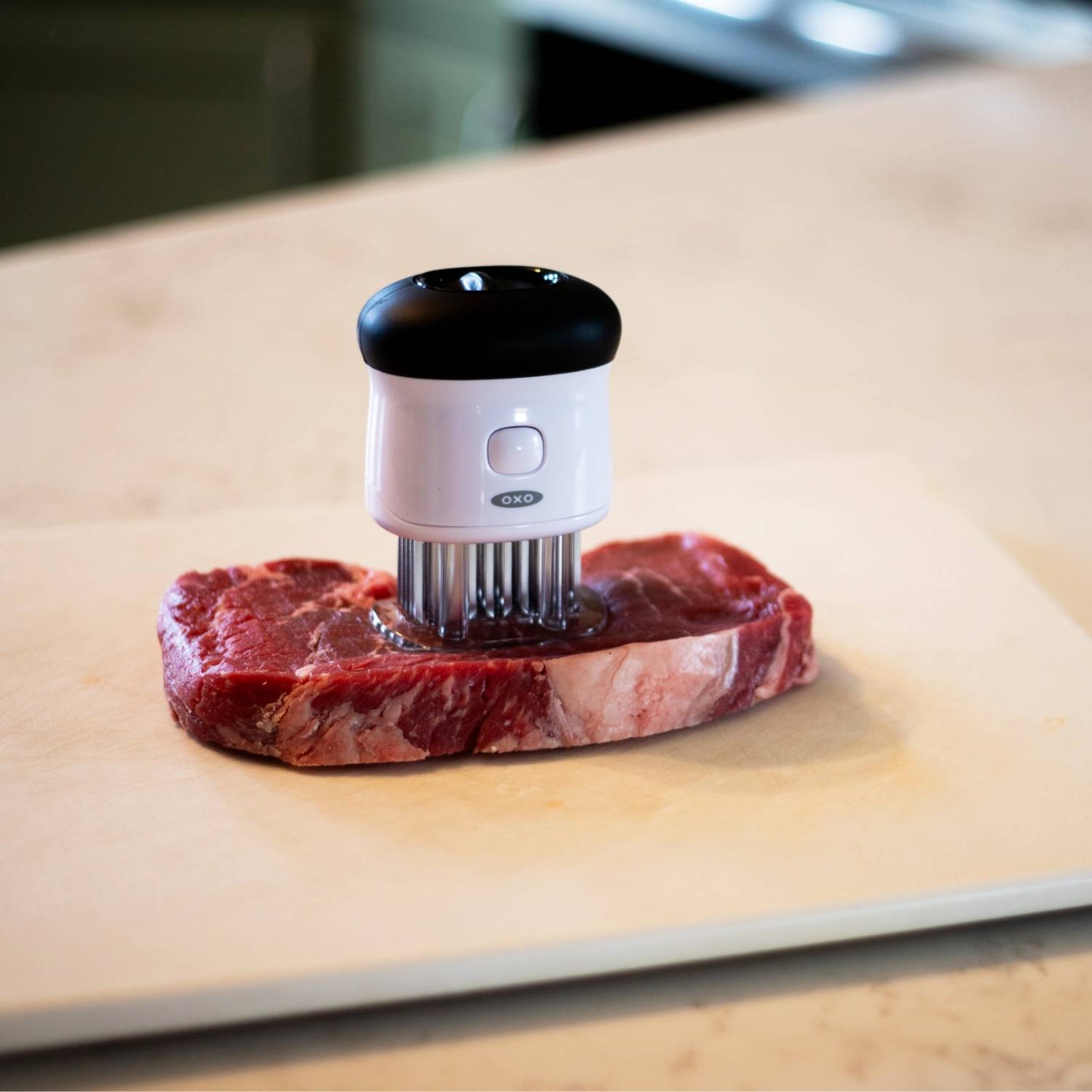Compact white and black meat tenderizer with 58 stainless steel blades inserted into a raw steak on a cutting board, part of Kitchen & Food, Specialty Equipment, Butchering Supplies, and Best Sellers collections.