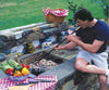 Person using a black powder-coated steel fire blower with brass mouthpiece and two-pronged claw to tend coals in a stone outdoor barbecue, surrounded by fresh vegetables and picnic items, ideal for Hearth Accessories and Stoves collections.