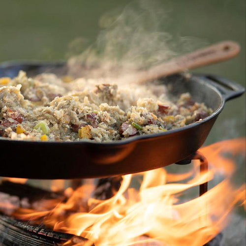 Handcrafted cast iron skillet with two side handles cooking a steaming savory dish over an open campfire, ideal for farm, garden, camping, kitchen, cookware, and cast iron use.
