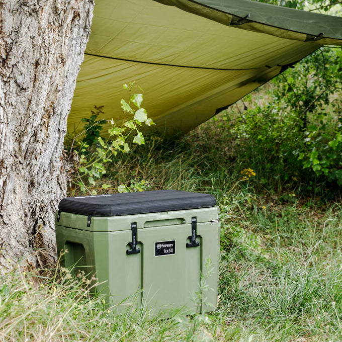 Olive green cooler box with black cushioned lid placed on grass beside a tree under a green tarp, suited for camping, farm, garden, and outdoor use in Best Sellers and Promo Eligible collections.