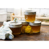 Four clear quilted glass jelly jars with metal lids filled with golden jam, arranged on a wooden surface with yellow flowers and a kitchen towel, for food preservation and canning.