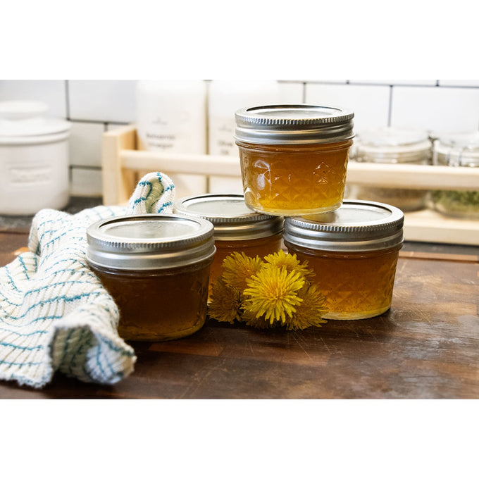 Four clear quilted glass jelly jars with metal lids filled with golden jam, arranged on a wooden surface with yellow flowers and a kitchen towel, for food preservation and canning.