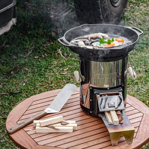 Cast iron skillet with two side handles cooking eggs and mushrooms over a wood-burning stove on a wooden table, ideal for farm, camping, kitchen, and cookware collections.